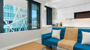 A modern living room with a blue sofa, a glass coffee table, and a large window with a geometric view. - Sleek 1 Bedroom Apartment (Cape Town)