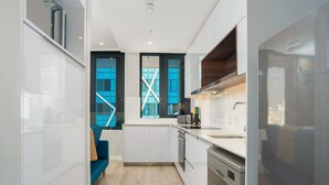 A compact kitchen with white cabinets, a stainless steel refrigerator, and a blue sofa in the adjacent living area. - Sleek 1 Bedroom Apartment (Cape Town)