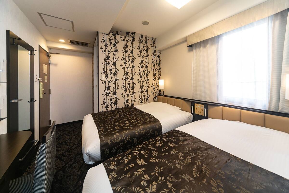 A hotel room with two beds, a desk, a chair, a window with curtains, and a wall with a floral pattern.