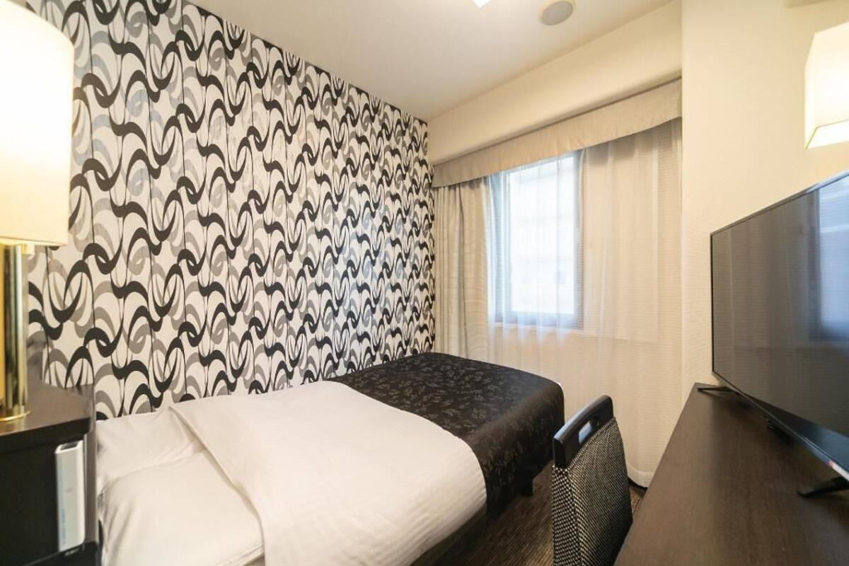 A hotel room with a bed, a TV, a desk, and a patterned wall.