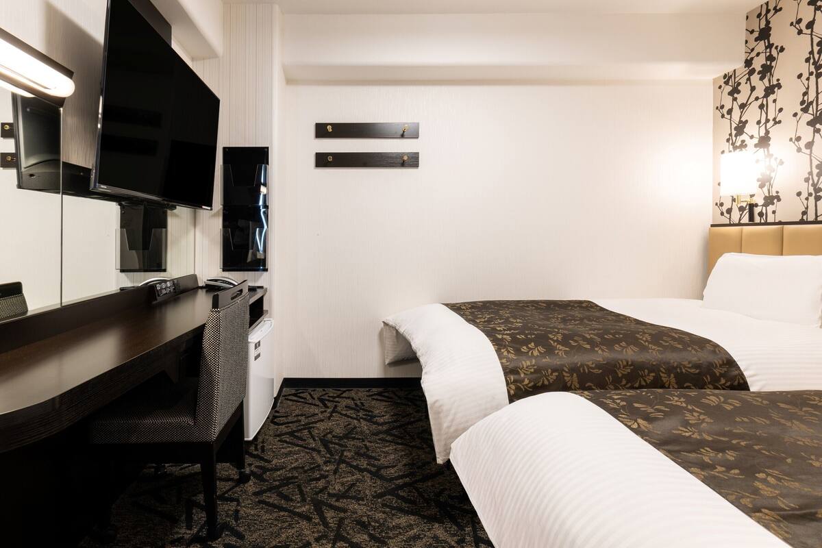 A hotel room with a desk, two beds, a flat-screen TV, and a wall-mounted TV.