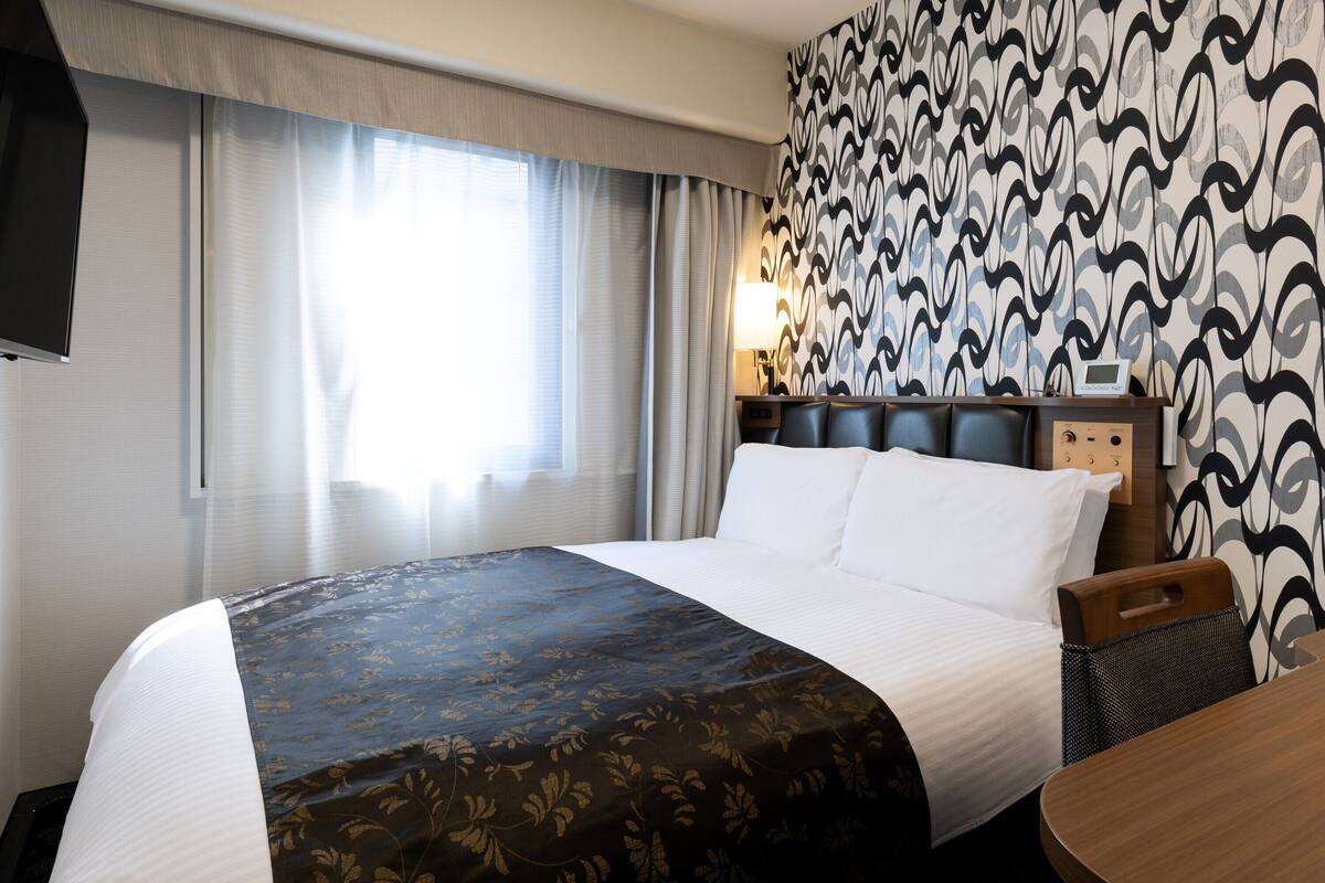 A hotel room with a large bed, a bedside table with a lamp, a TV mounted on the wall, and a patterned wallpaper.