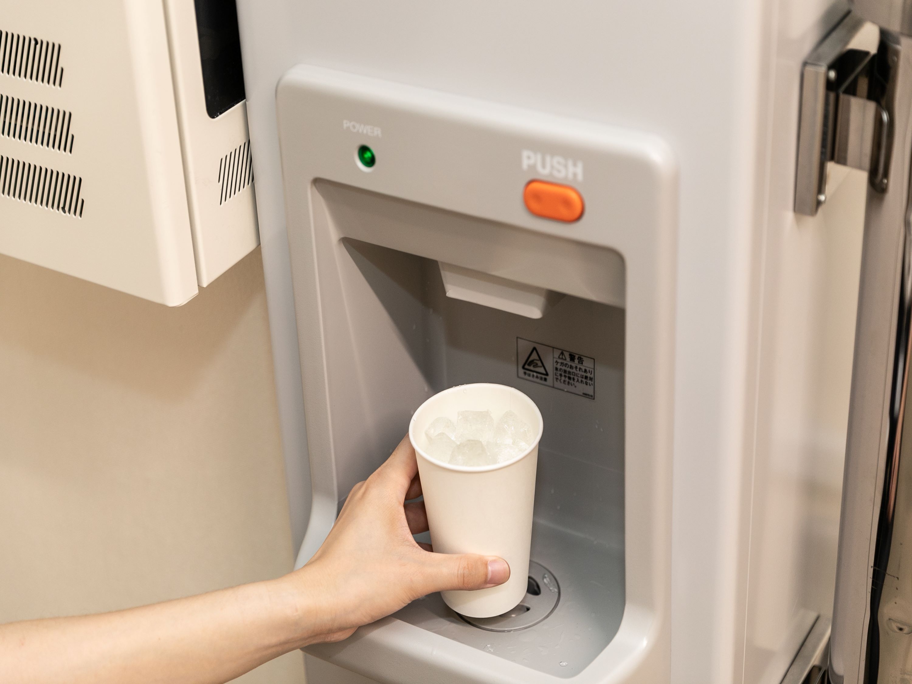 A hand holding a cup at a water dispenser with a green power button and an orange push button.