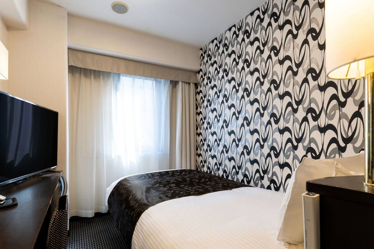 A hotel room with a bed, a TV, a desk, and a patterned wall.