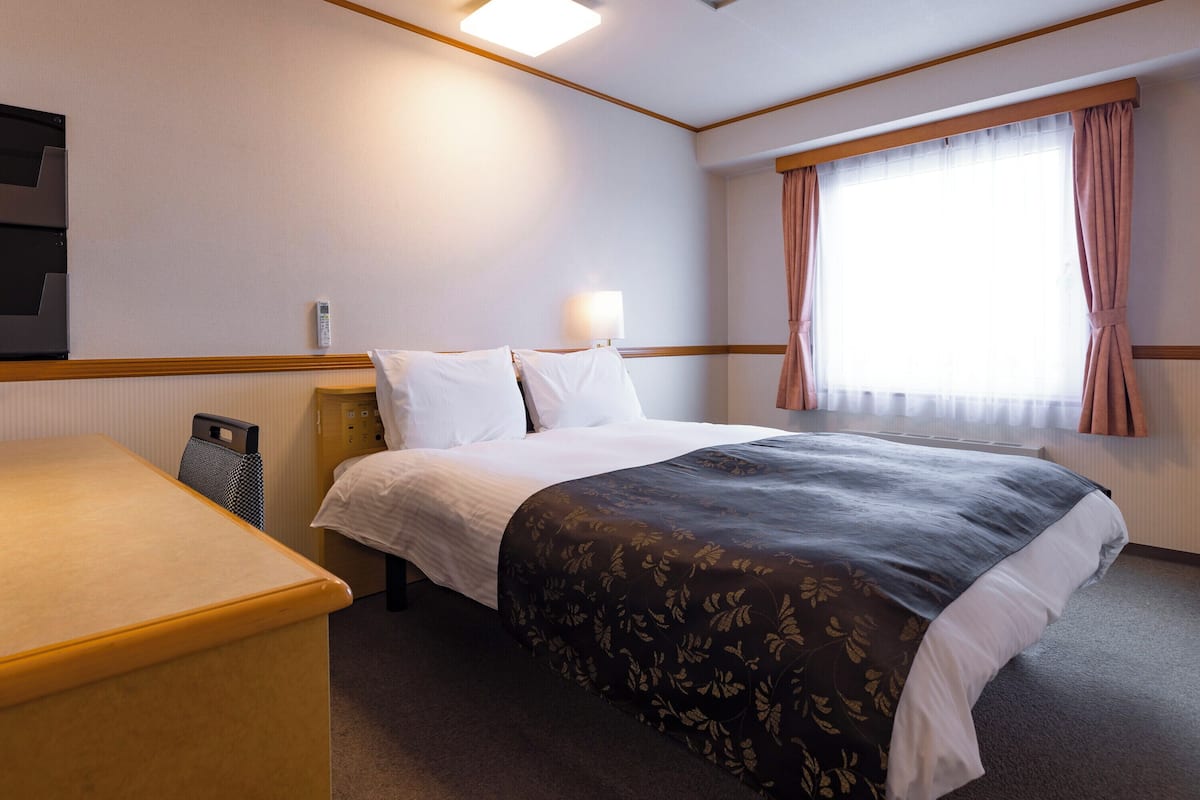 A hotel room with a bed, bedside table, lamp, and a window with curtains.