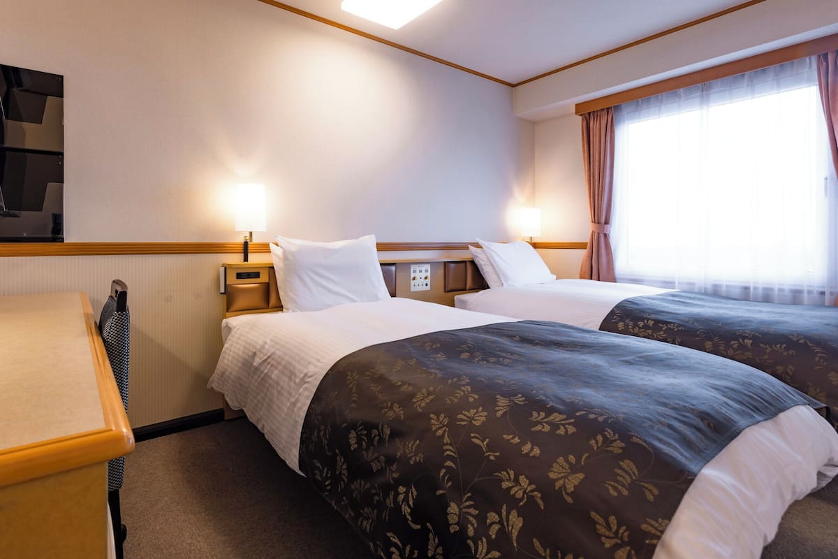 A hotel room with two beds, a desk, a TV, and a window with curtains.