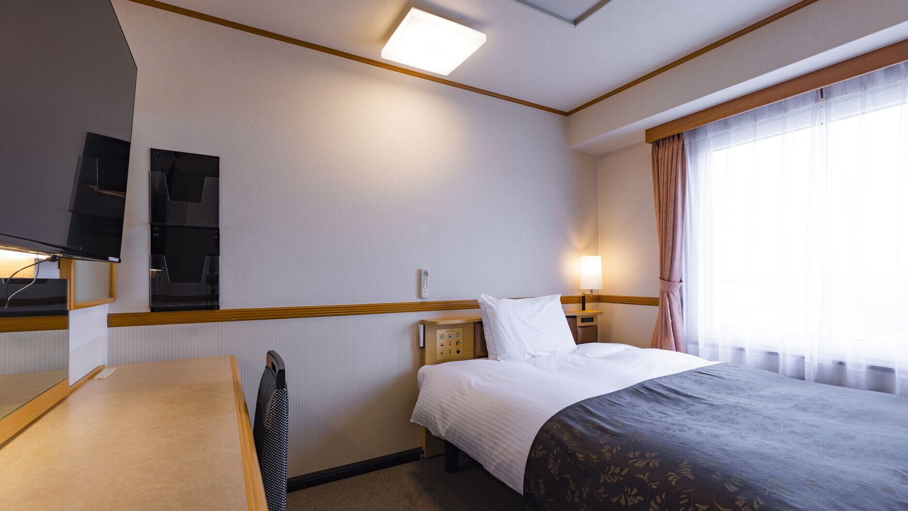Hotelkamer met een bed, nachtkastje, lamp, televisie en een raam met gordijnen.
