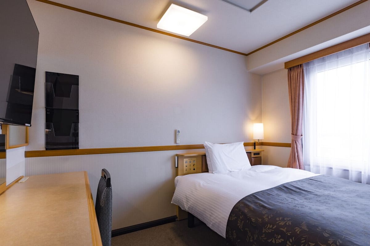 A hotel room with a bed, bedside table, lamp, TV, and a window with curtains.