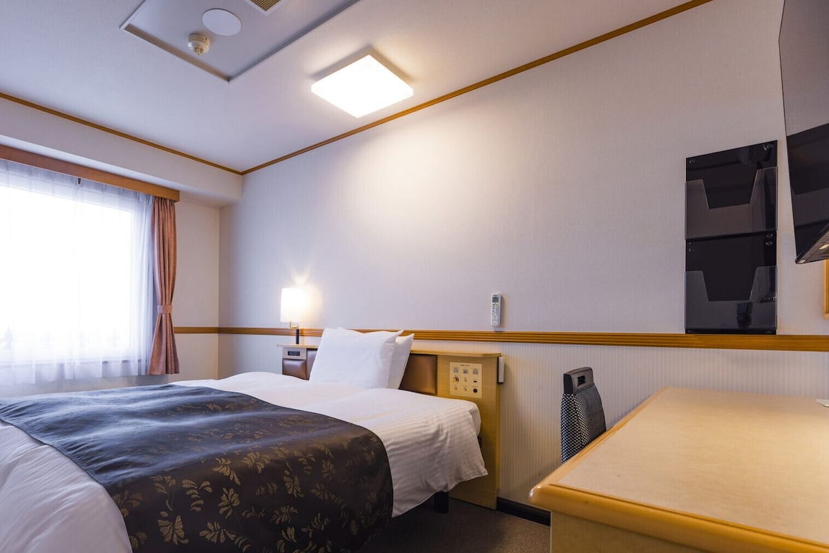 A hotel room with a bed, bedside table, lamp, and a small desk.