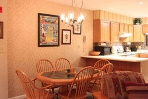 A dining area with a round table and six chairs, a chandelier, and a kitchen with wooden cabinets and a tiled backsplash.