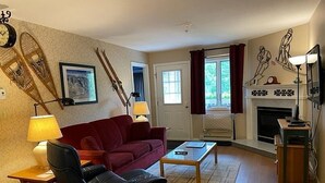Un salon avec un canapé rouge, un fauteuil noir, une table basse en bois et une cheminée. On y trouve deux lampes sur des tables d’appoint, une horloge au mur et un tableau encadré au-dessus du canapé.