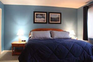 Ein Schlafzimmer mit blauer Wand, einem Bett mit blauer Bettdecke, zwei gerahmten Bildern an der Wand und einem Nachttisch mit Lampe.