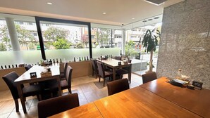 A modern restaurant with wooden tables and chairs, large windows offering a view of the city, and a stone accent wall. - APA Hotel Keikyukamata Ekimae (Tokyo)