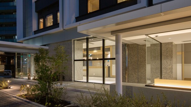 A modern building with large glass windows, a well-lit entrance, and a landscaped outdoor area with plants. - APA Hotel Keikyukamata Ekimae (Tokyo)