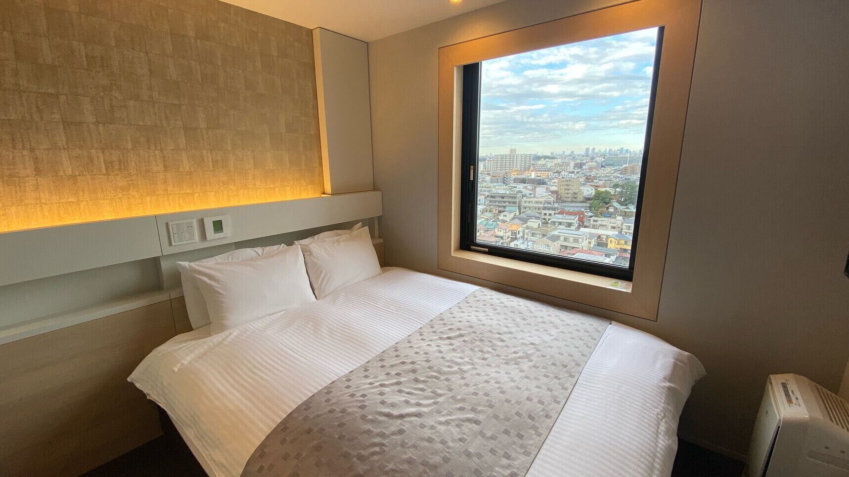 Une chambre d’hôtel avec un lit, une fenêtre donnant sur la ville, une lampe fixée au mur et un radiateur.