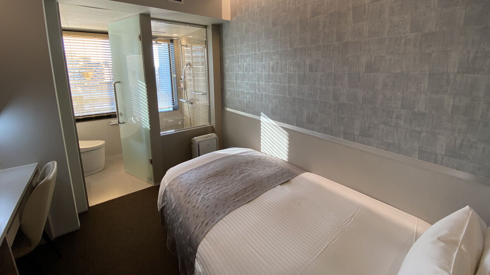 Une chambre d’hôtel comprenant un lit, un bureau, une chaise, une fenêtre avec des stores et un espace douche.