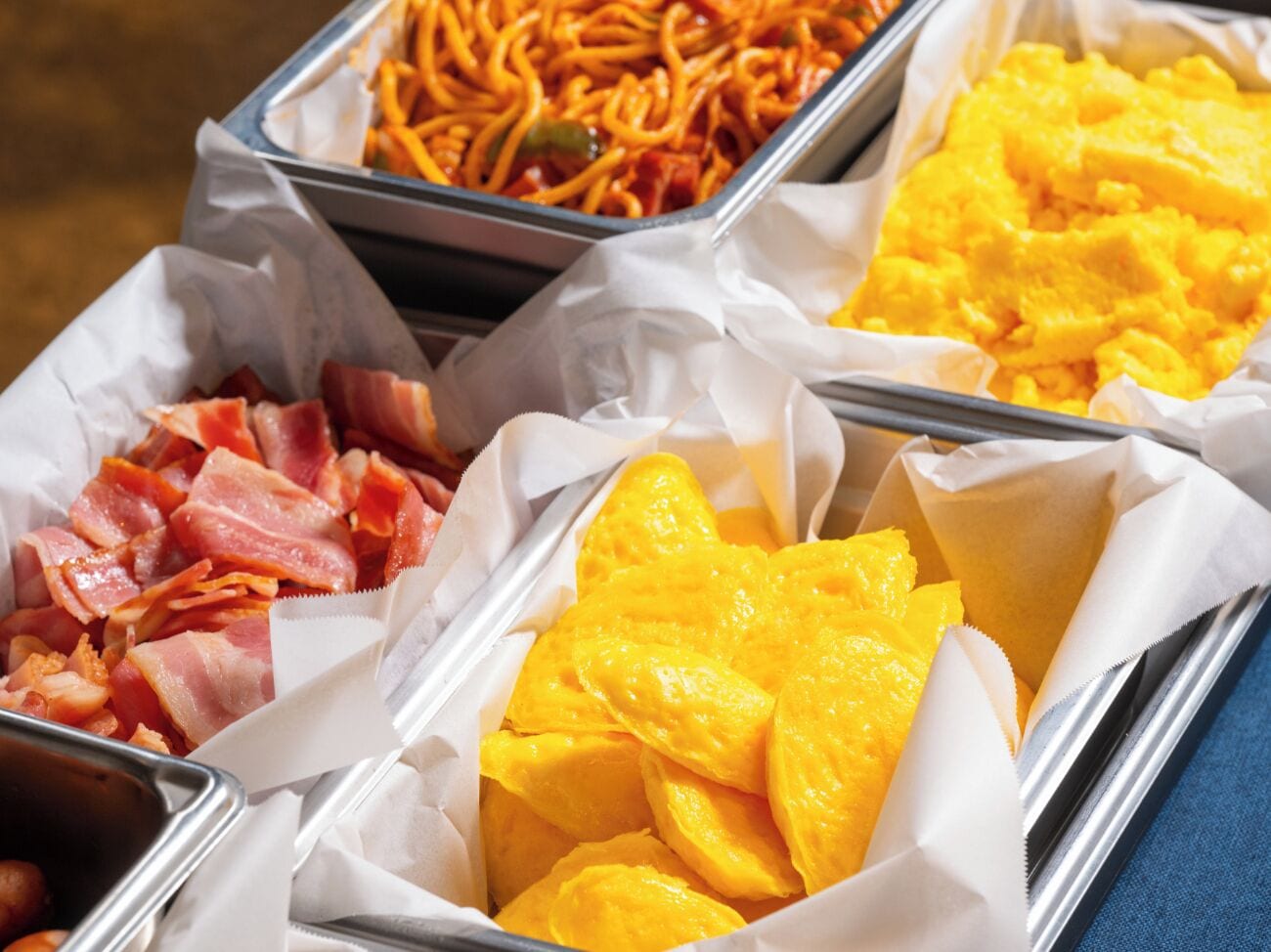 Un bufé con distintos alimentos, entre ellos, tocino, fideos y gajos de mango.