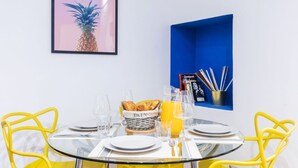 Ett matbord med gula stolar, en glasskiva och en korg med bröd. På väggen hänger en inramad bild av en ananas, och det finns en blå hylla med böcker och en kopp med pennor.