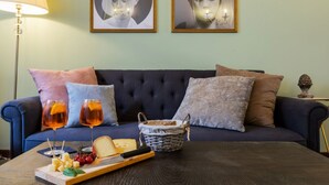 Ein Wohnzimmer mit einem dunkelblauen Sofa, einem Holztisch mit einer Käseplatte, Weingläsern mit Rosé und einem Korb mit dem Aufdruck "PAIN".