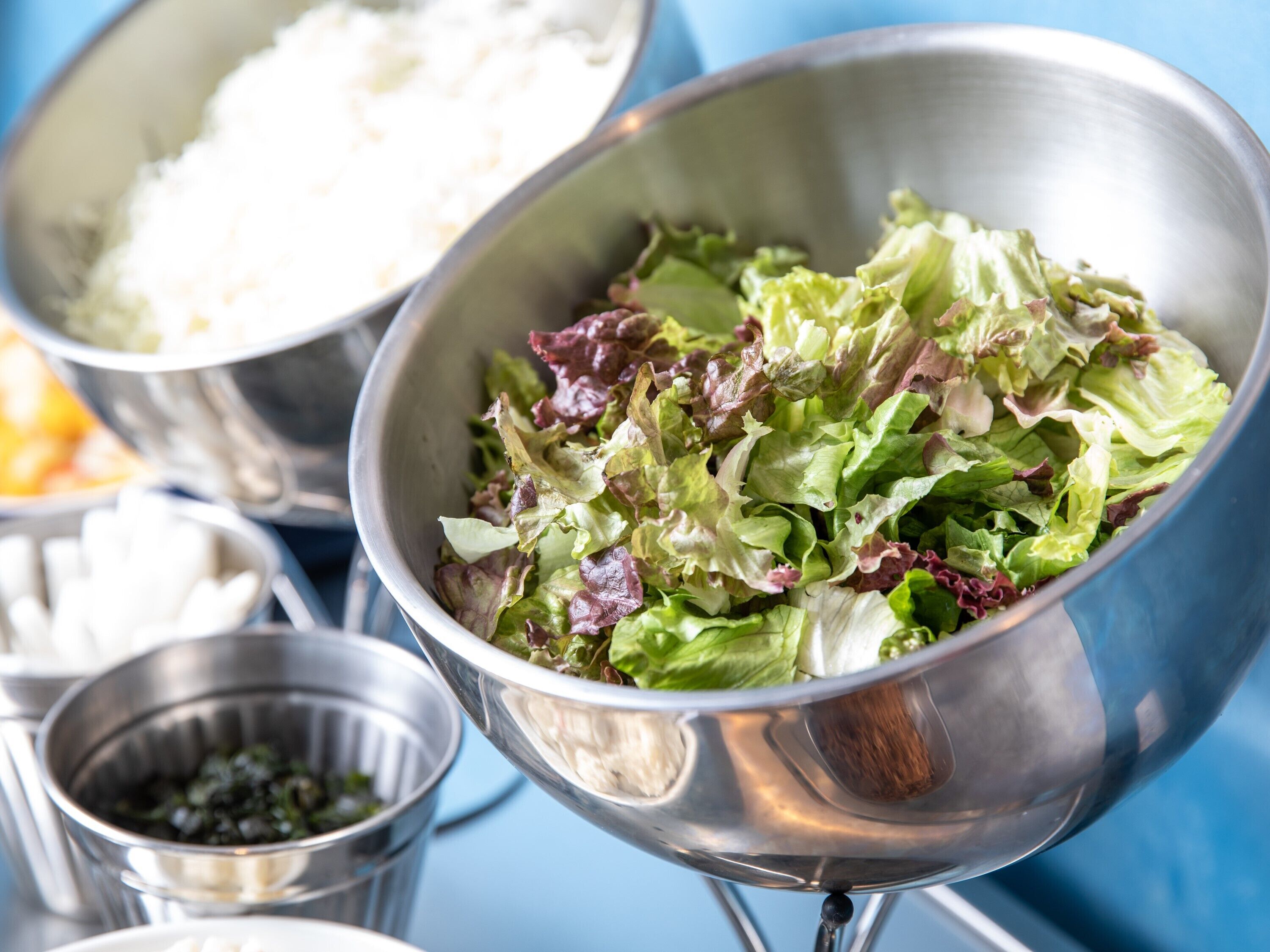 A metal bowl filled with mixed green and red lettuce leaves, surrounded by other metal bowls containing white rice and green herbs.