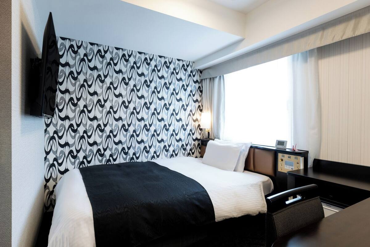 A modern hotel room with a large bed, a bedside table with a lamp, and a view of the outdoors through a window.