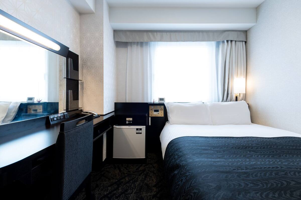 A hotel room with a bed, a desk, a chair, a small refrigerator, a mirror, and a window with curtains.