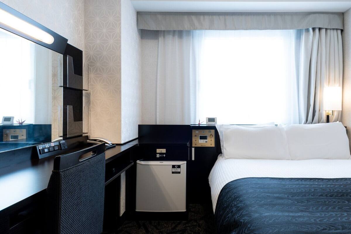 A hotel room with a bed, a desk, a chair, a small fridge, a bedside table, a lamp, and a window with curtains.
