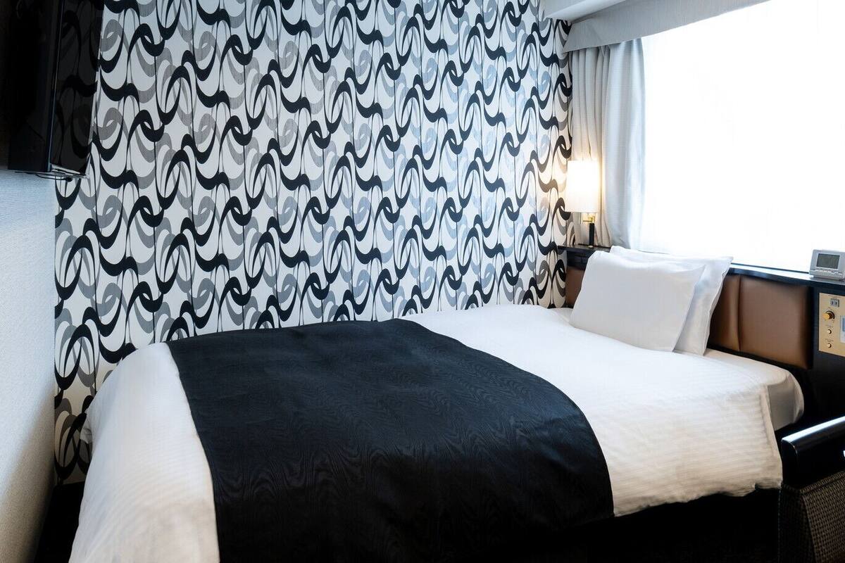 A hotel room with a bed, a nightstand, a lamp, and a control panel.