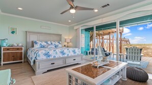 Quarto com cama, mesa de cabeceira, penteadeira, ventilador de teto e vista para a praia através de uma porta de vidro.