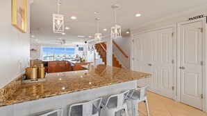 Cozinha com bancadas de granito, banquetas brancas, iluminação pendente e vista para o exterior.