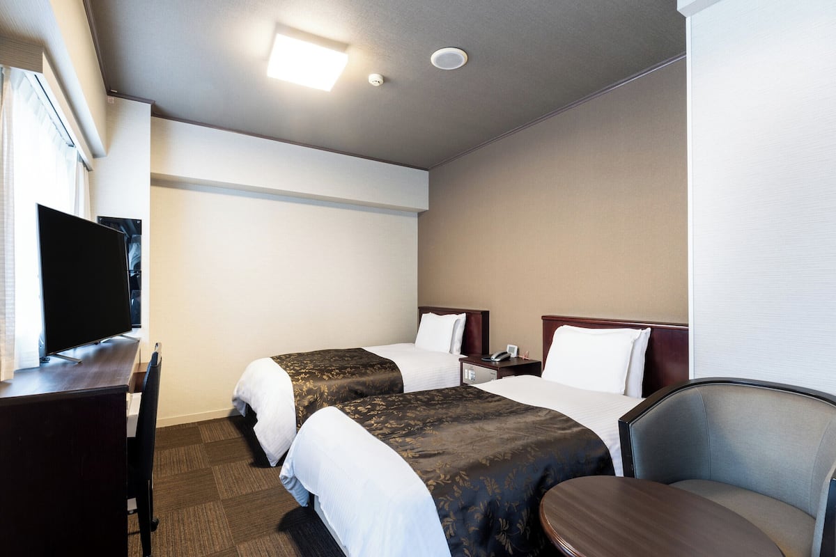 Quarto de hotel com duas camas, uma escrivaninha, televisão de tela plana, uma cadeira e uma mesinha.