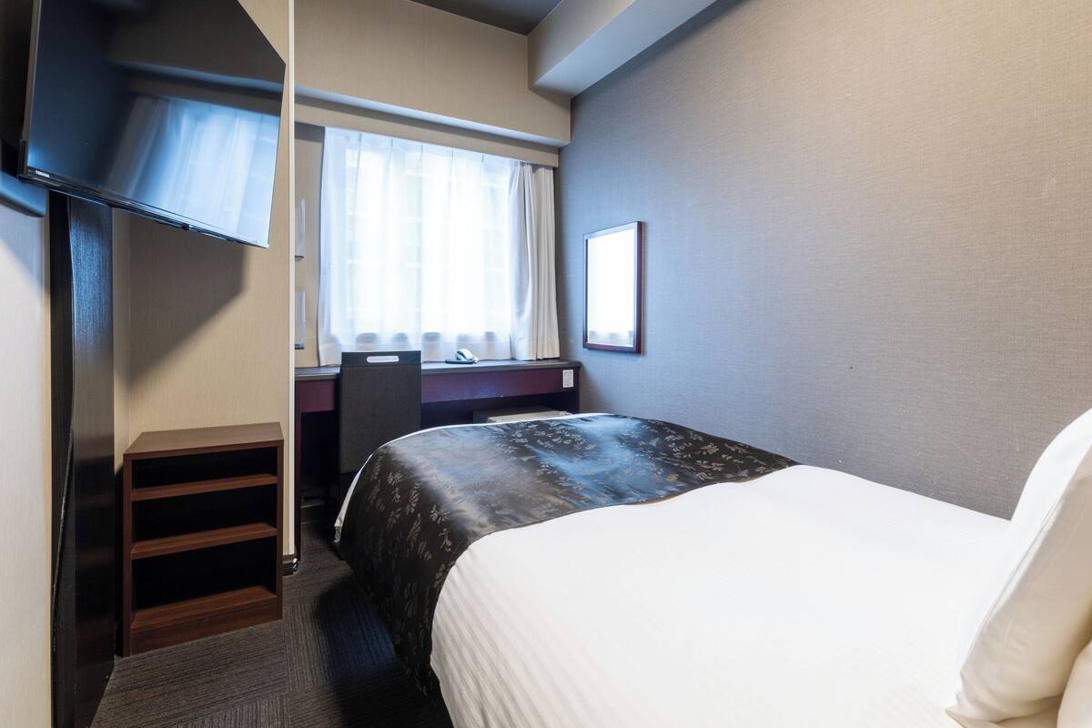 Quarto de hotel com cama, escrivaninha, cadeira, televisão e janela com cortinas.