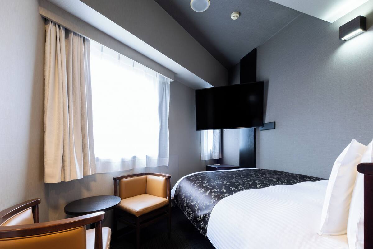Quarto de hotel com cama, uma cadeira, uma mesinha, televisão fixada na parede e uma janela com cortinas.