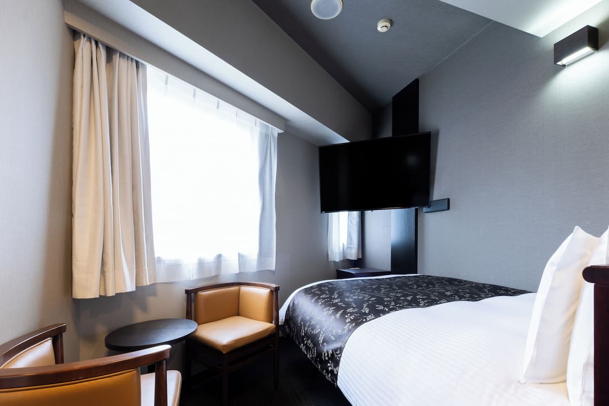 A hotel room with a bed, a chair, a small table, a TV mounted on the wall, and a window with curtains.