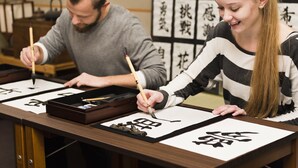 Two individuals practicing calligraphy at a wooden table with black ink and brushes. - APA Hotel Asakusa Kaminarimon Minami (Tokyo)