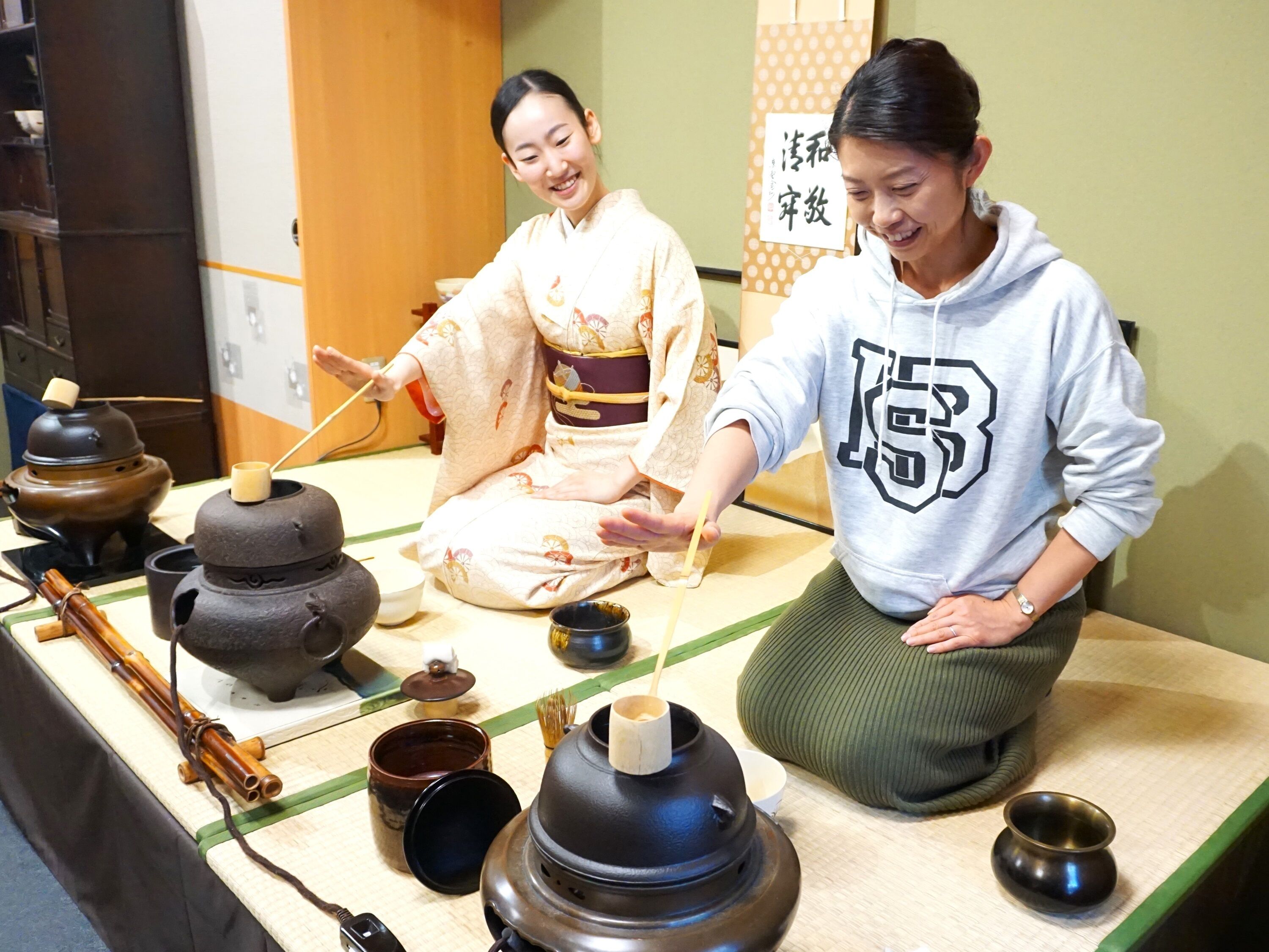 Dos personas sentadas a una mesa con utensilios para una ceremonia tradicional del té japonesa, incluyendo tazas de té y un batidor de bambú.