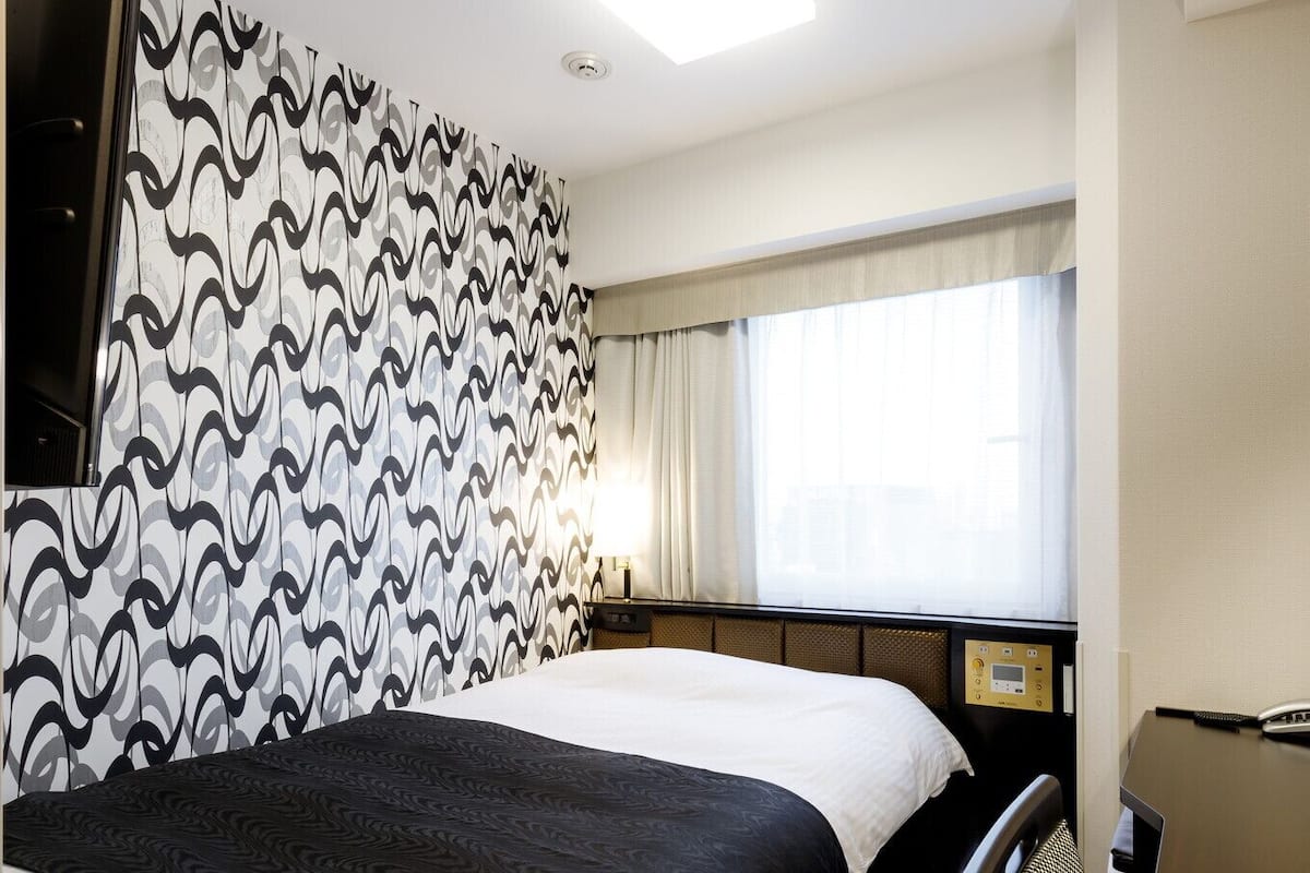 Habitación de hotel con cama, escritorio con teléfono, ventana con cortinas y pared con papel tapiz estampado.