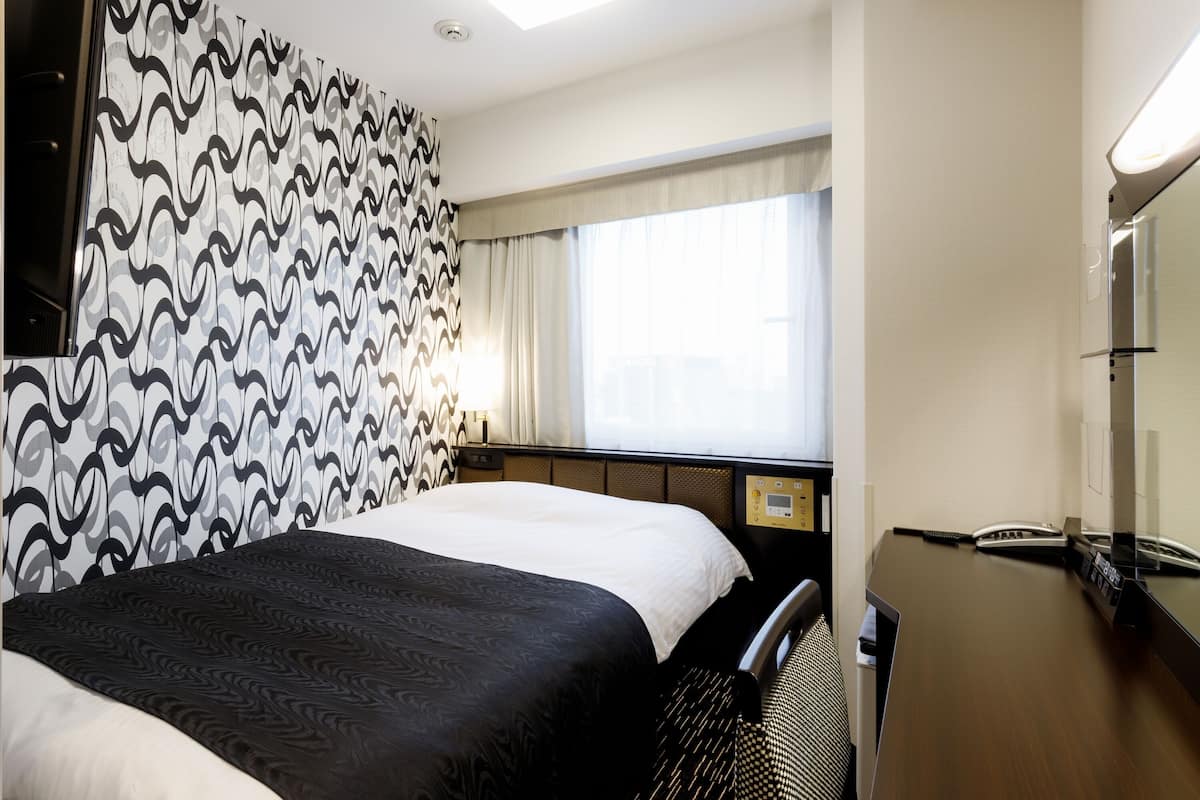 Habitación de hotel con cama, escritorio, silla, ventana con cortinas y pared con papel tapiz estampado.