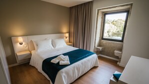 Un lit bien fait, avec du linge de lit blanc et un plaid turquoise, une lampe de chevet et une fenêtre donnant sur des arbres.