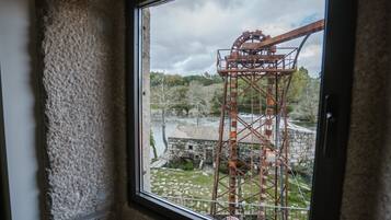 A window view of a large industrial water wheel structure with a stone building and greenery in the background.