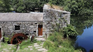 Un bâtiment rustique en pierre, doté d’une grande roue à eau, d’une porte en bois et d’un sentier de pierre menant à un plan d’eau.