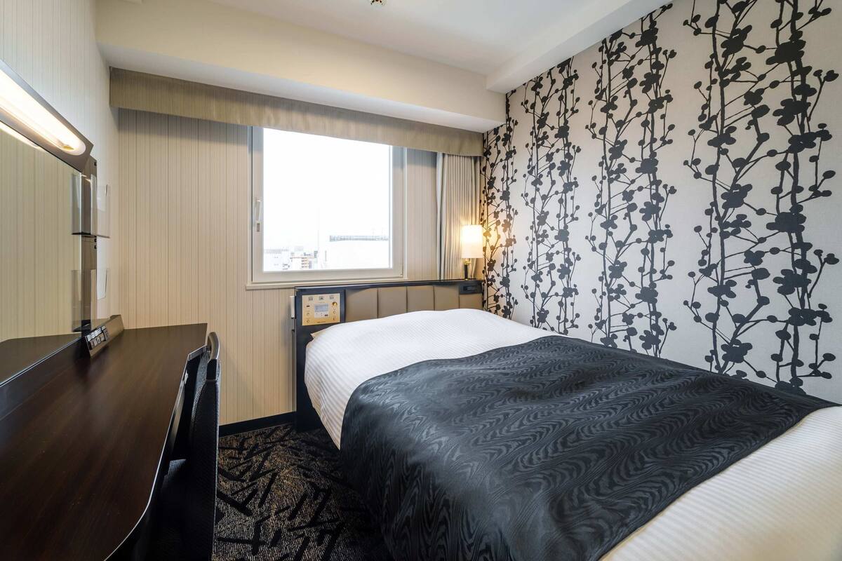 Ein Hotelzimmer mit Bett, Nachttisch, Lampe, Fenster und Wand mit Blumenmuster.