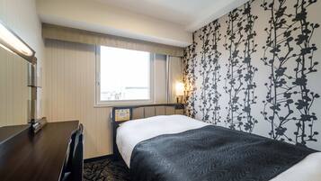 A hotel room with a bed, bedside table, lamp, window, and a wall with a floral pattern.