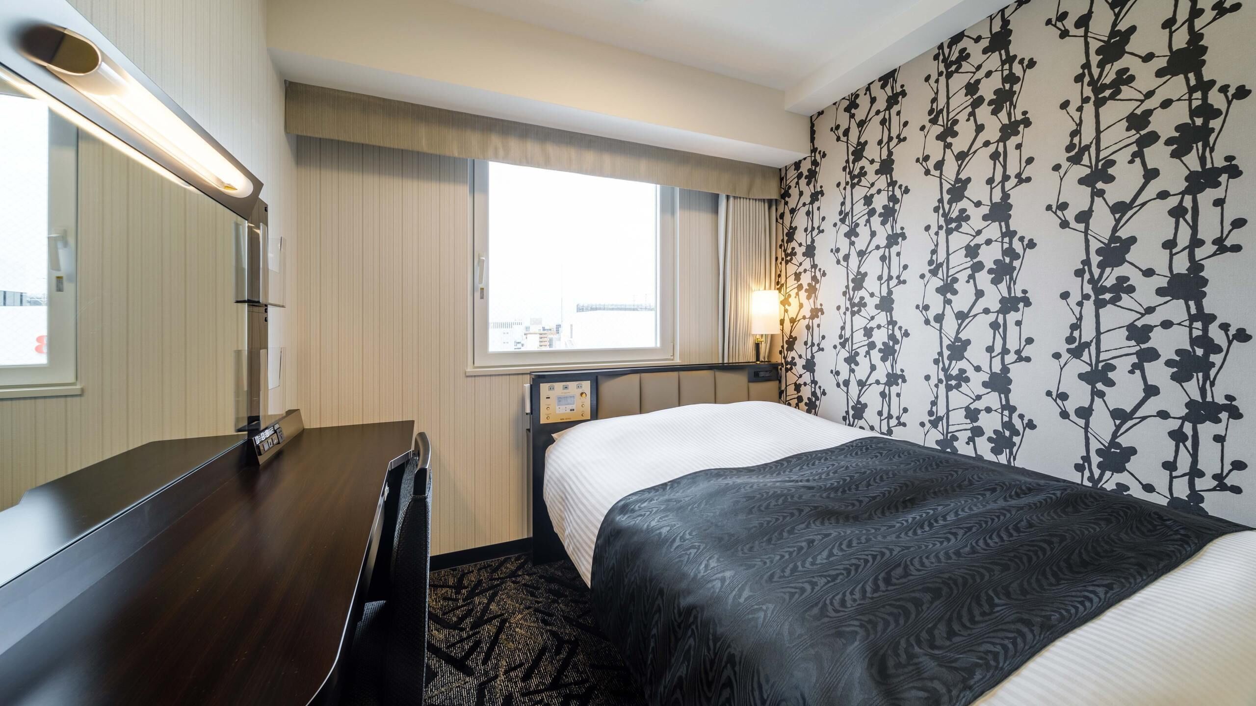 Ein Hotelzimmer mit Bett, Nachttisch, Lampe, Fenster und einer Wand mit Blumenmuster.