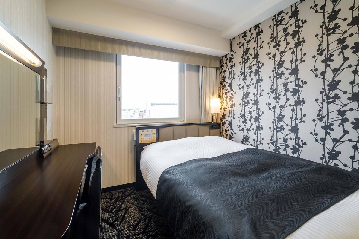 A hotel room with a bed, bedside table, lamp, window, and a wall with a floral pattern.