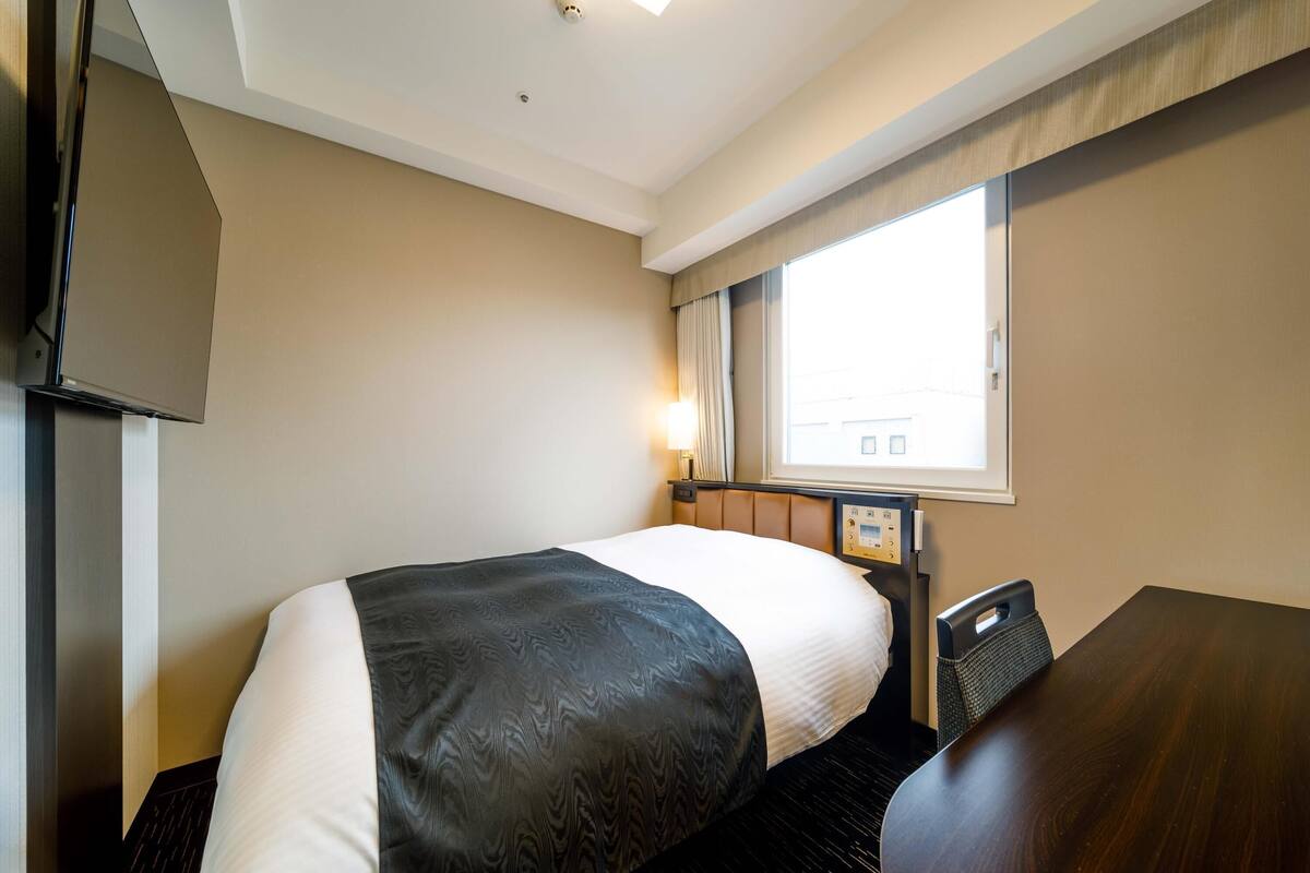 Ein Hotelzimmer mit Bett, Schreibtisch, Fenster und Bedienfeld.