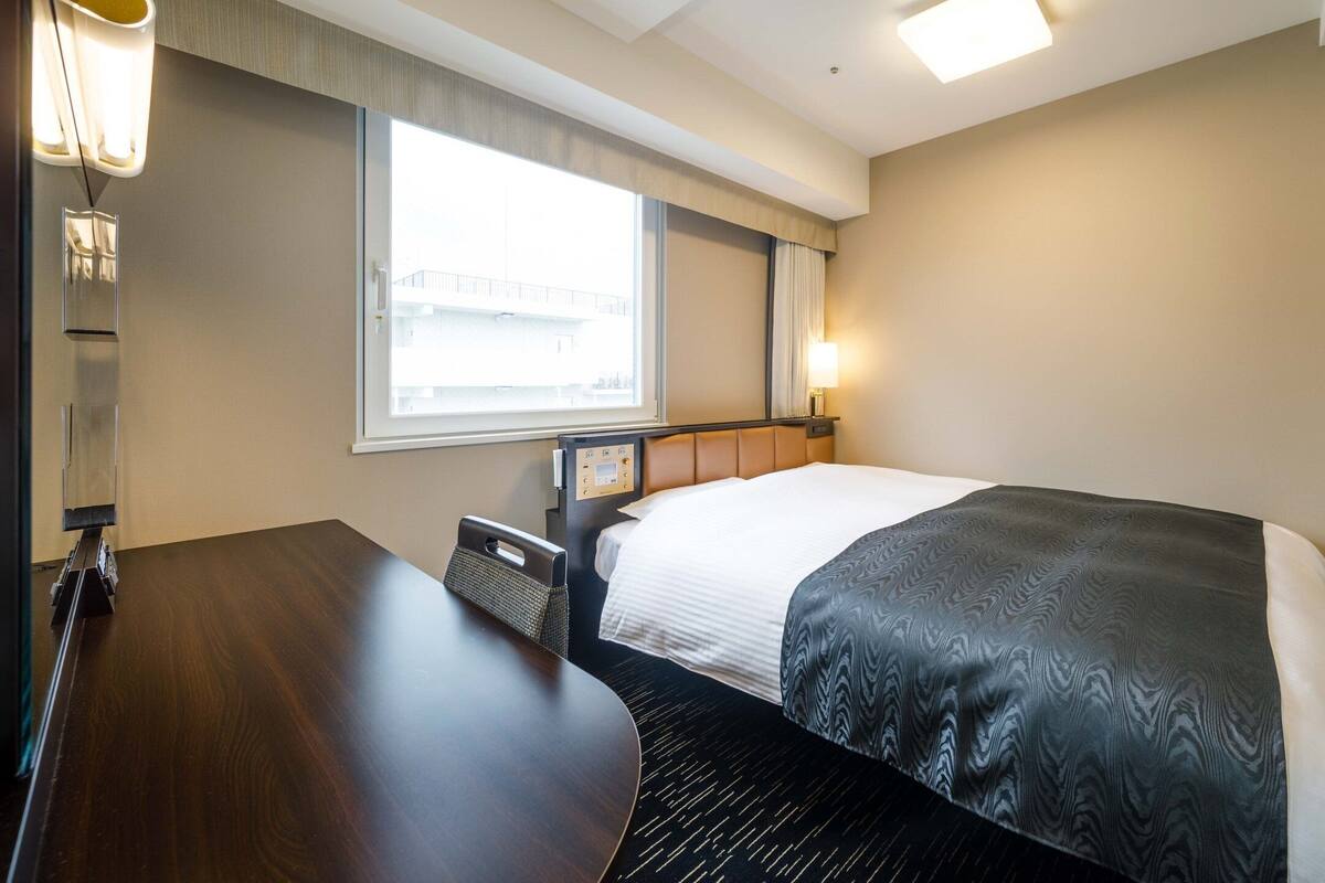 Ein Hotelzimmer mit Bett, Nachttisch, Lampe, Fenster und Bedienfeld.