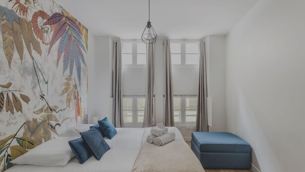 Un dormitorio con una cama, un banco azul, un aplique colgante y un tapiz floral grande.