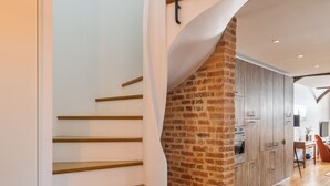 Un escalier moderne avec des marches en bois et un mur d’accent en briques, menant à un espace cuisine doté de meubles en bois et d’une table à manger.