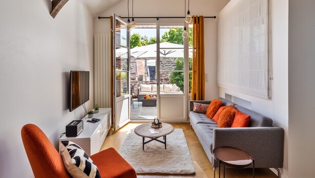 Un salon moderne avec un canapé, une table basse et un téléviseur. Une grande fenêtre, ornée de rideaux, offre une vue sur l’extérieur.